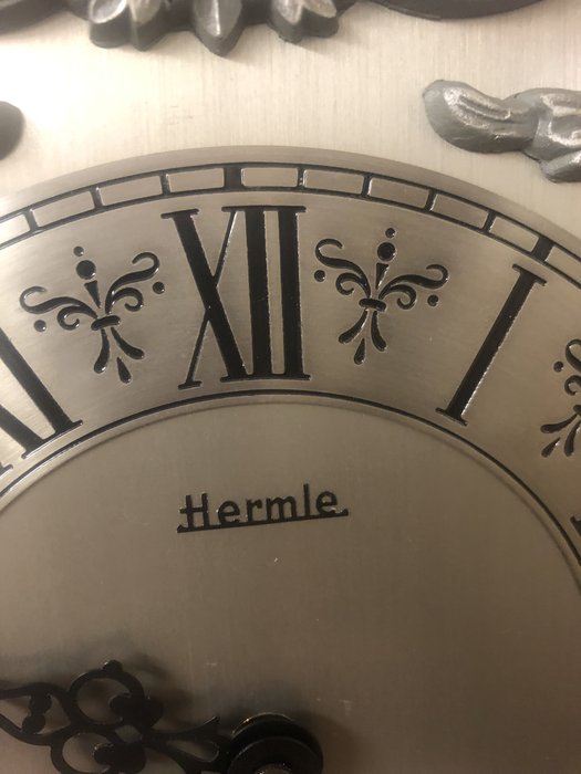 Nástěnné hodiny, vyrobeny  německou firmou  Hermle. Elegantní ciferník, kolem něhož jsou okrasné motivy. 